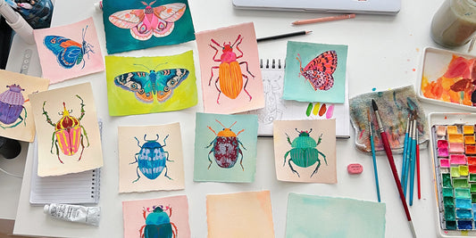Flat lay of hand-painted beetle and butterfly illustrations on textured watercolor paper.