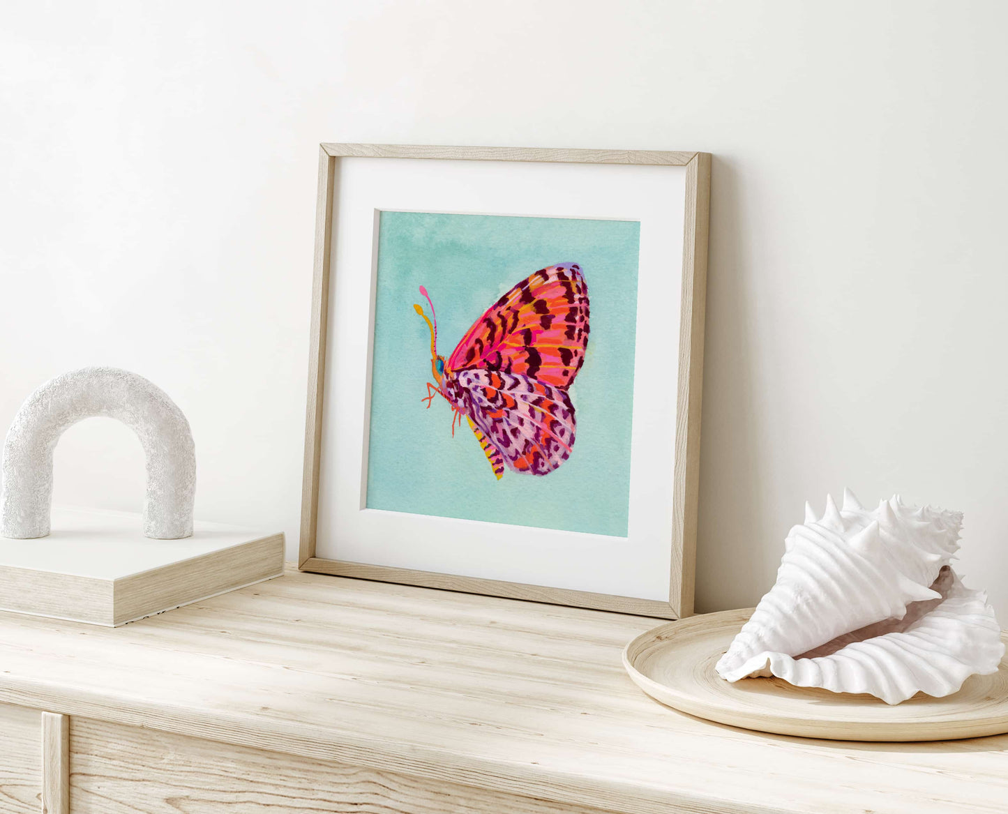 Framed artwork of a colorful butterfly on a light wooden surface with decorative shells.