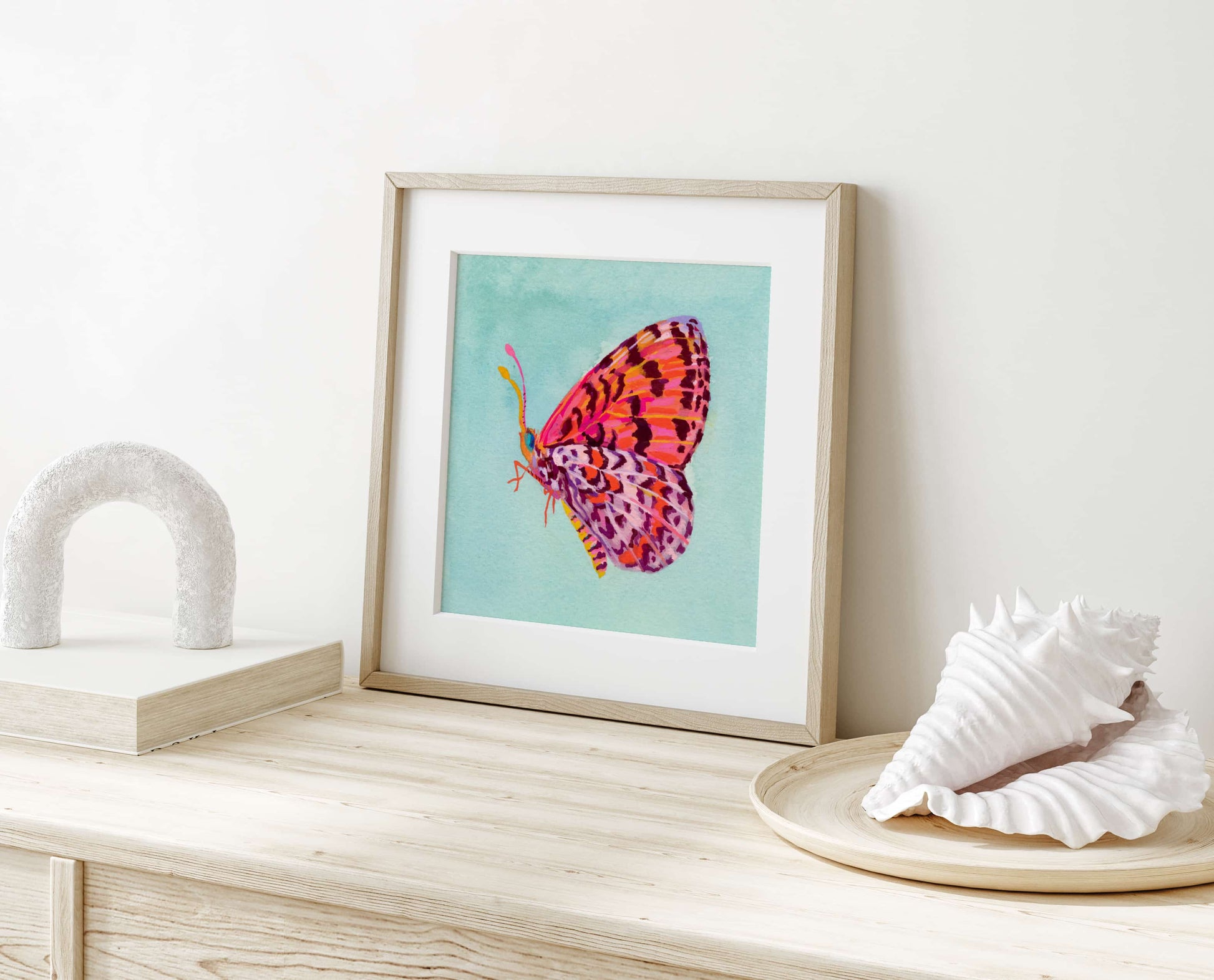 Framed artwork of a colorful butterfly on a light wooden surface with decorative shells.