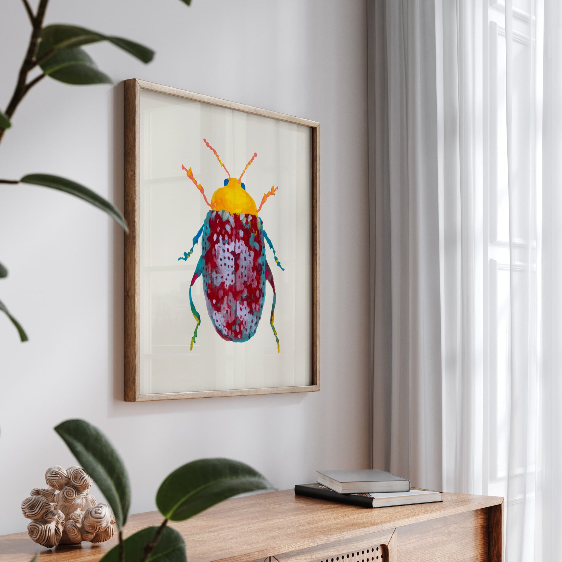 Colorful beetle artwork in a wooden frame on a wall above a wooden surface with books and a plant.